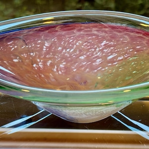 May include: A shallow, oval-shaped glass bowl featuring a translucent, multi-colored design. The interior displays a textured, web-like pattern in shades of pink and orange, while the outer rim is accented with a vibrant green glass border.