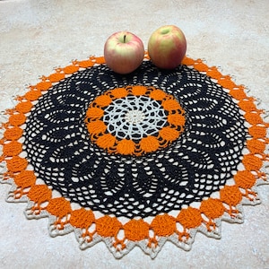 May include: A black and orange crocheted doily with a white center. Two red apples are sitting on top of the doily.