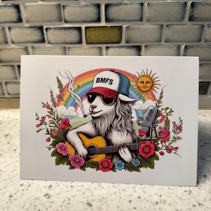 May include: A white greeting card featuring a cartoon goat wearing sunglasses and a hat with "BMFS" on it, playing a yellow guitar. The card has a rainbow, sun, microphone, and floral accents.