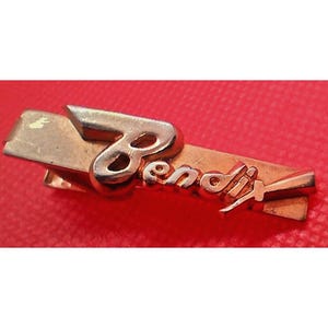 May include: A gold-coloured tie clip featuring the name "Bendix" in a cursive script. The clip has a rectangular form with a raised, stylised "B" at the top. The background is a solid red colour.