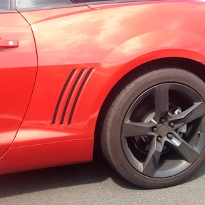 Peut inclure: Une voiture de sport rouge avec des accents noirs sur l'aile arrière et des roues noires. La voiture est garée sur une surface d'asphalte grise.