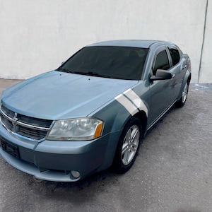 Puede incluir: Un sedán Dodge Avenger azul claro con rayas de carreras blancas en el lateral. El coche tiene ventanas tintadas y una parrilla cromada.