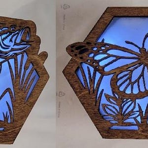 Peut inclure: Deux boîtes lumineuses décoratives en bois de forme hexagonale. L'une présente un motif de poisson, l'autre un papillon. Les deux ont une lumière bleue qui brille à travers les motifs découpés, créant un effet lumineux. Le bois a une teinte chaude naturelle.