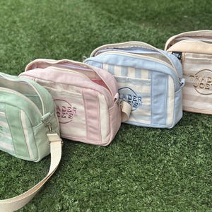 May include: Four rectangular crossbody bags in pastel green, pink, light blue, and beige. Each bag features a striped pattern, a front pocket, a zip closure, an adjustable strap, and the embroidered text Trader Joe's in a circular logo.