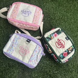 May include: Three rectangular crossbody bags with adjustable straps. One bag features pink floral panels and purple embroidery. Another has lavender trim and floral accents. The third bag displays a dark green border with a tropical hibiscus print.