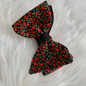 Può includere: Un papillon nero con un motivo floreale rosso. Il papillon è realizzato in tessuto e presenta un piccolo motivo a foglie verdi sottile.