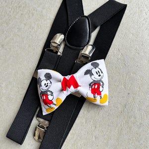 May include: Black suspenders with a white bow tie featuring a Mickey Mouse print. The bow tie has red accents and yellow shoes on the Mickey Mouse characters.