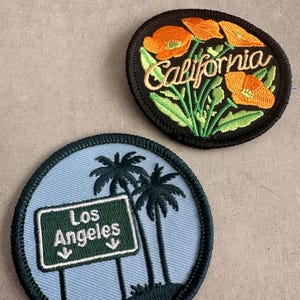 May include: Two embroidered patches. One features orange California poppies with the word "California" in cursive on a black background. The other patch has a light blue background with a green sign that reads "Los Angeles" and two palm trees.