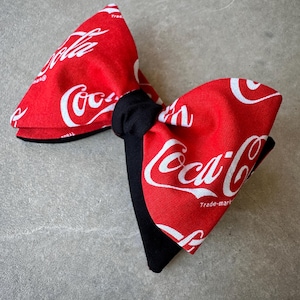 May include: Red bow tie with a white Coca-Cola logo pattern. The bow tie is tied with a black ribbon.