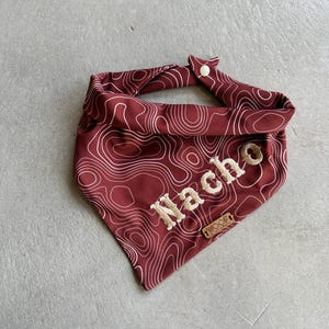 Op de afbeelding: Een rode bandana met een wit topografisch patroon en het woord "Nacho" in wit geborduurd. De bandana heeft een klein bruin leren label met het woord "Wanderlust" erop.
