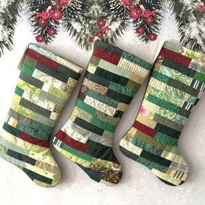 May include: Three handmade Christmas stockings with a patchwork design in shades of green, red, and cream. The stockings are decorated with various patterns and textures. Festive holiday decor.
