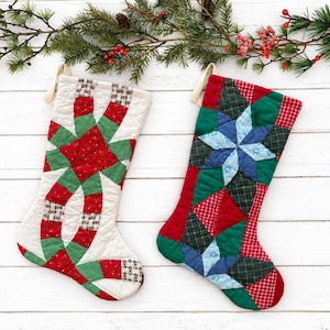 Peut inclure: Deux chaussettes de Noël matelassées avec des motifs en patchwork complexes. La chaussette de gauche présente un motif entrelacé rouge et vert sur fond blanc. La chaussette de droite arbore un motif en étoile multicolore rouge, vert et bleu.