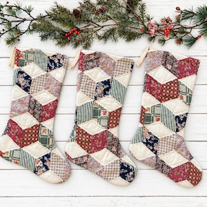 May include: Three quilted Christmas stockings featuring a geometric patchwork design. Each stocking is constructed from various fabric squares in shades of red, blue, green, and cream, accented with floral and striped patterns and a hanging loop.
