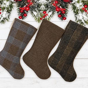 May include: Three Christmas stockings arranged side by side. The left stocking features a grey and brown plaid pattern. The middle stocking is a solid brown textured fabric. The right stocking displays a dark brown and black glen plaid pattern.