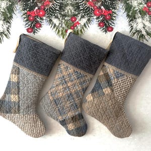 May include: Three quilted Christmas stockings with grey tops and patterned bodies in shades of blue, tan, and grey. The stockings feature various geometric and plaid patterns. Festive holiday decor.