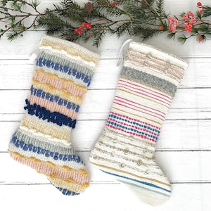 May include: Two woven Christmas stockings with textured, colorful designs. One features blue, yellow, and pink stripes with a dark blue pom-pom accent. The other has pink, gray, and gold stripes. Both have white hanging loops.