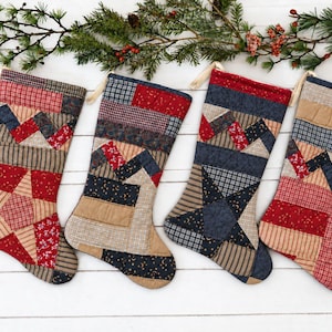 May include: Four quilted Christmas stockings featuring patchwork designs in red, navy blue, tan, and cream fabrics. Each stocking displays a unique arrangement of geometric patterns, including stars and stripes, with a hanging loop at the top.