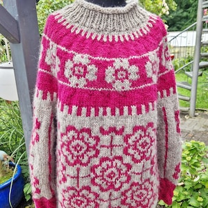 Könnte beinhalten: Ein langärmeliger Strickpullover in leuchtendem Fuchsia-Pink und Haferbeige. Das Kleidungsstück zeigt ein sich wiederholendes Blumenmuster auf der Brust und dem Körper, abgerundet mit einem gerippten Stehkragen und gerippten Bündchen.