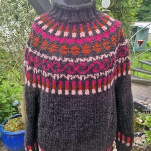 Könnte beinhalten: Ein dunkelgrauer, handgestrickter Pullover mit hohem Kragen. Der Schulterbereich weist ein lebendiges Muster aus rosa, orangefarbenen und weißen geometrischen Designs auf. Die Manschetten und der Saum haben ebenfalls ein passendes rosa-rotes Muster. Dieser gemütliche Pullover ist perfekt für kaltes Wetter.