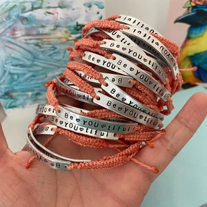 K&ouml;nnte beinhalten: Ein Stapel handgefertigter Armb&auml;nder mit korallenfarbenen gewebten B&auml;ndern. Jede silberfarbene Metallplatte ist mit den Worten "Be YOUtiful" und einem Herzsymbol versehen. Die Armb&auml;nder werden in einer Hand gehalten.