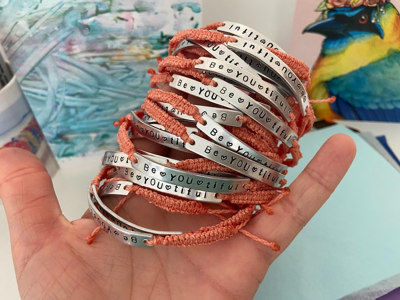 K&ouml;nnte beinhalten: Ein Stapel handgefertigter Armb&auml;nder mit korallenfarbenen gewebten B&auml;ndern. Jede silberfarbene Metallplatte ist mit den Worten "Be YOUtiful" und einem Herzsymbol versehen. Die Armb&auml;nder werden in einer Hand gehalten.
