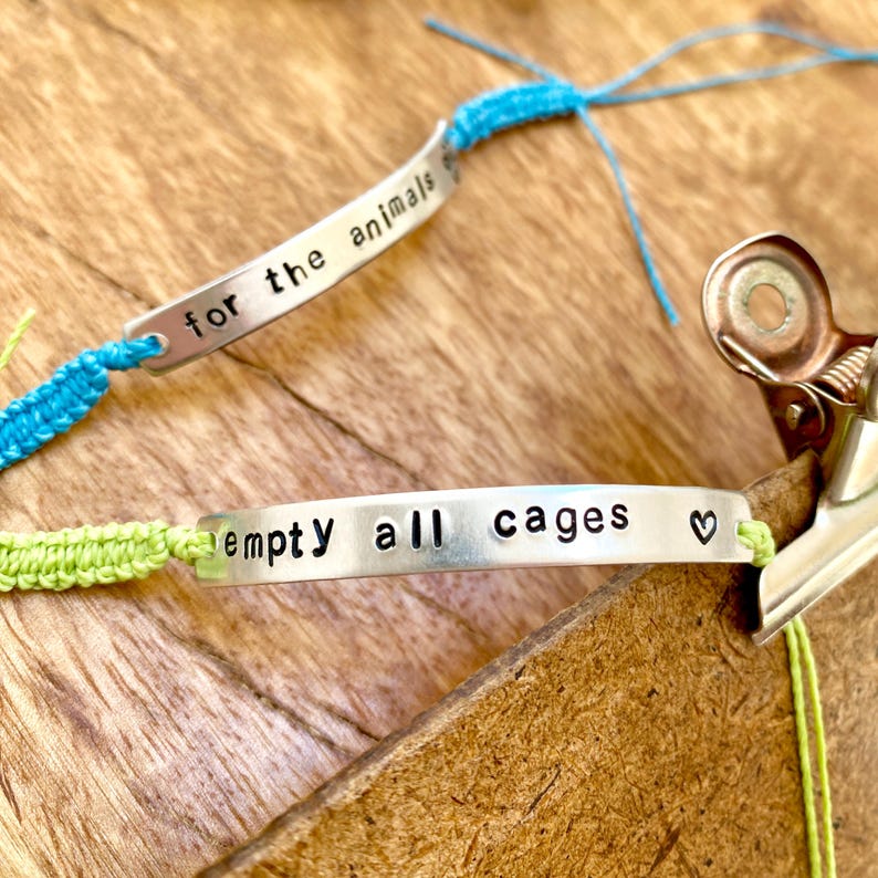 A crafting scene featuring two silver-toned metal bar bracelets with black stamped text. One bracelet features the phrase "for the animals" on a bright blue cord, while the other displays the text "empty all cages" with a small heart symbol on a lime green cord.