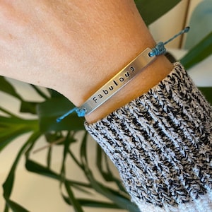 K&ouml;nnte beinhalten: Ein silbernes Armband mit dem Wort "Fabulous" eingestanzt. Das Armband ist an einer blauen Kordel befestigt und wird am Handgelenk getragen. Die Person tr&auml;gt einen schwarz-wei&szlig; gestreiften Pullover.