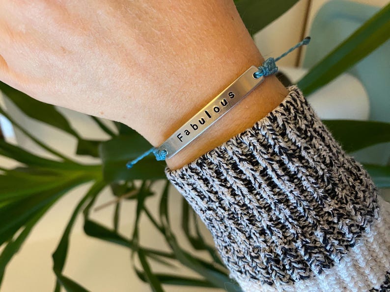 K&ouml;nnte beinhalten: Ein silbernes Armband mit dem Wort "Fabulous" eingestanzt. Das Armband ist an einer blauen Kordel befestigt und wird am Handgelenk getragen. Die Person tr&auml;gt einen schwarz-wei&szlig; gestreiften Pullover.