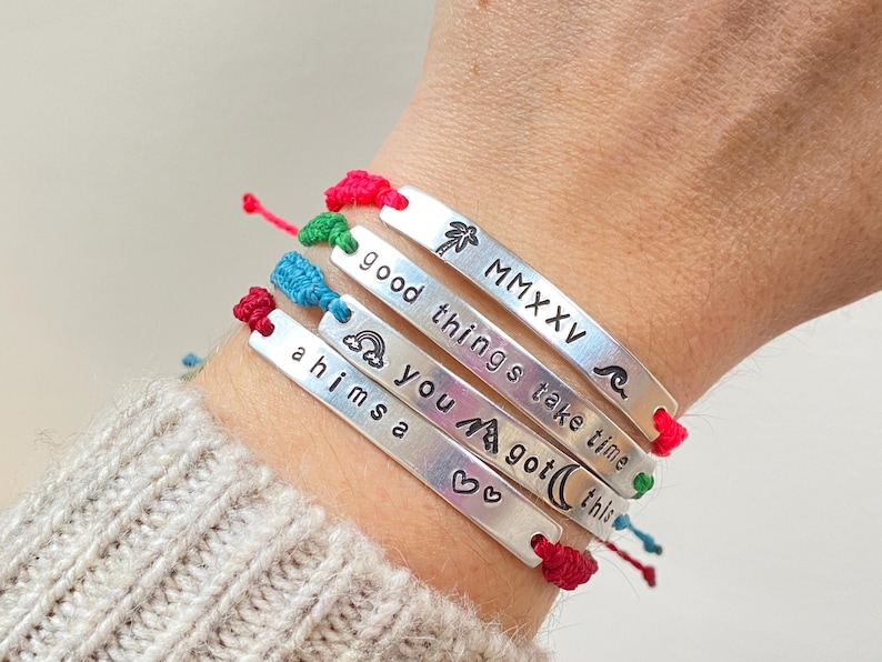 A set of custom engraved bar bracelets featuring braided cord wristbands are worn on a persons wrist. Each bracelet has a stamped personalisation of words, phrases, dates and symbols on a silver-coloured metal bar. The wristbands are made from braided cord and are multicoloured. The bracelets shown are stamped with phrases "good things take time", "ahimsa" and "MMXV" Roman numerals. They feature engravings of the emojis of a palm tree, a wave, a mountain and a rainbow.