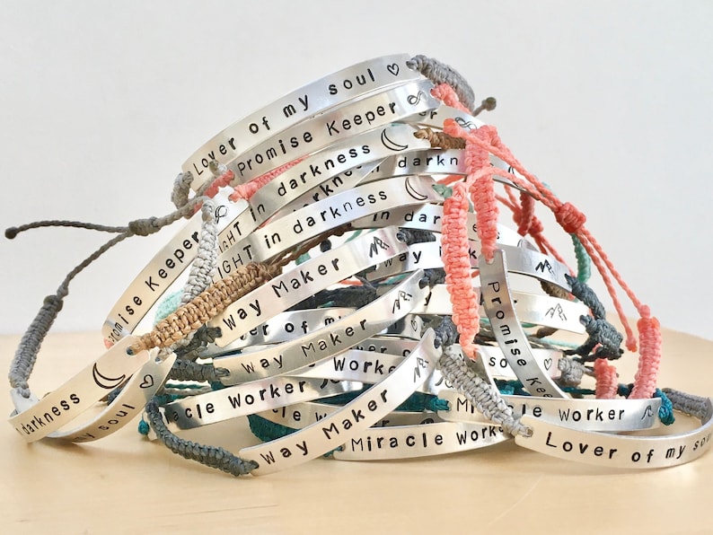 a large order off handmade metal silver bar bracelets. bracelets stamped with motivational text. each custom made with a personalised engraving and has a woven adjustable wristband in mixed colours. bracelets customised with emojis.
