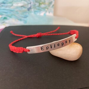 K&ouml;nnte beinhalten: Ein rotes Makramee-Armband mit einem silbernen Balken, auf dem das Wort "Epilepsy" steht. Das Armband liegt auf einem kleinen, glatten, hellfarbigen Stein. Der Hintergrund ist ein verschwommener Farbmix.
