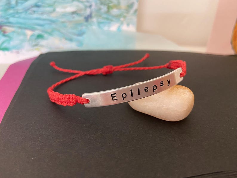 K&ouml;nnte beinhalten: Ein rotes Makramee-Armband mit einem silbernen Balken, auf dem das Wort "Epilepsy" steht. Das Armband liegt auf einem kleinen, glatten, hellfarbigen Stein. Der Hintergrund ist ein verschwommener Farbmix.