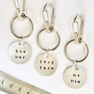 May include: Three silver keychains with metal rings and clips. Each keychain has a round metal tag with the words "She Her", "They Them", and "He Him" engraved on them.