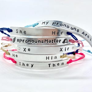 Peut inclure: Une pile de bracelets en argent avec des cordons colorés. Chaque bracelet porte un ensemble de pronoms différent gravé dessus, notamment "Elle/Elle", "Il/Il", "Iel/Iel", "Xe/Xir" et "#PronounsMatter".