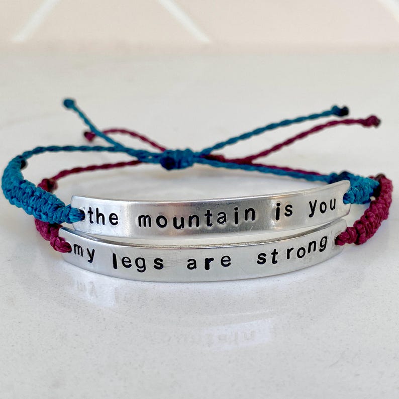 Two silver-toned metal bar bracelets with black stamped text. The top bar reads "the mountain is you" and the bottom bar reads "my legs are strong". The bracelets feature adjustable blue and maroon braided cord bands.