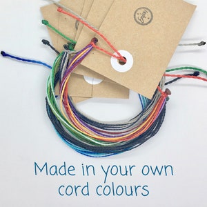 May include: A selection of multi-coloured cord bracelets with brown tags. The bracelets are in various colours, including blue, green, red, and grey. The text "Made in your own cord colours" is written in blue.
