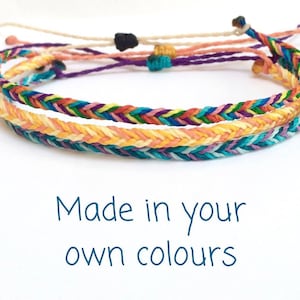 Könnte beinhalten: Mehrfarbiges geflochtenes Freundschaftsarmband mit Chevron-Muster. Das Armband ist aus verschiedenen Farben gefertigt, darunter Gelb, Orange, Rosa, Blau, Grün und Lila. "Made in your own colours" steht unter dem Armband.