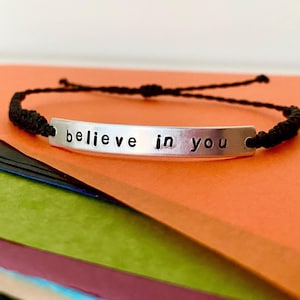 K&ouml;nnte beinhalten: Ein silbernes Armband mit dem Schriftzug "believe in you". Das Armband hat ein schwarzes geflochtenes Kordelband. Der Hintergrund ist eine Mischung aus Orange, Gr&uuml;n und Schwarz.