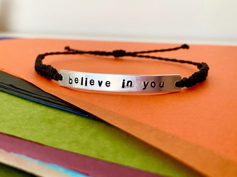 K&ouml;nnte beinhalten: Ein silbernes Armband mit dem Schriftzug "believe in you". Das Armband hat ein schwarzes geflochtenes Kordelband. Der Hintergrund ist eine Mischung aus Orange, Gr&uuml;n und Schwarz.