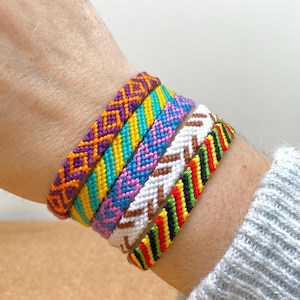 Op de afbeelding: Een verzameling van vijf kleurrijke vriendschapsarmbanden. De armbanden hebben ingewikkelde geweven patronen in verschillende kleuren, waaronder paars, oranje, geel, turquoise, roze, wit, bruin, rood, groen en zwart. De armbanden worden om een pols gedragen.