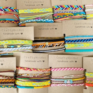 Może przedstawiać: Kolekcja kolorowych, ręcznie robionych bransoletek przyjaźni. Bransoletki mają różne wzory i kolory, w tym tęczowy, niebieski, żółty i różowy. Każda bransoletka jest prezentowana na brązowej karcie z napisem "CraftyMotMot handmade with" i symbolem serca.