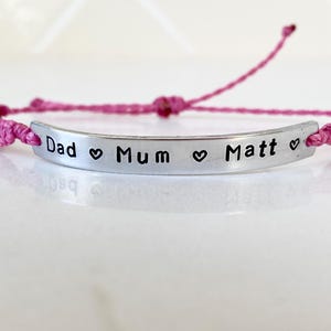K&ouml;nnte beinhalten: Ein rosa gewebtes Armband mit einem silbernen Balken. Auf dem Balken stehen die Worte "Dad", "Mum" und "Matt", getrennt durch Herzsymbole. Das Armband ist verstellbar.