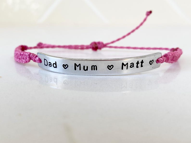 K&ouml;nnte beinhalten: Ein rosa gewebtes Armband mit einem silbernen Balken. Auf dem Balken stehen die Worte "Dad", "Mum" und "Matt", getrennt durch Herzsymbole. Das Armband ist verstellbar.