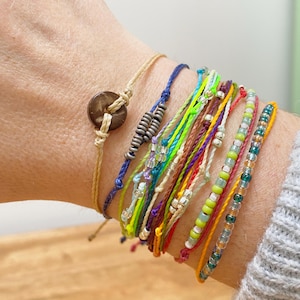 May include: A selection of vibrant bracelets are displayed on a wrist. The bracelets include a beige cord bracelet with a wooden button, a blue cord bracelet, and several beaded and woven bracelets in various colours like green, yellow, and red.