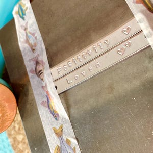 K&ouml;nnte beinhalten: Zwei silberne Metallarmb&auml;nder mit den Worten "Positivity" und "Loved" sind abgebildet. Kleine Herzformen sind neben den Worten gestempelt. Dekoratives Klebeband mit bunten Fischmotiven ist auf dem Metall angebracht.