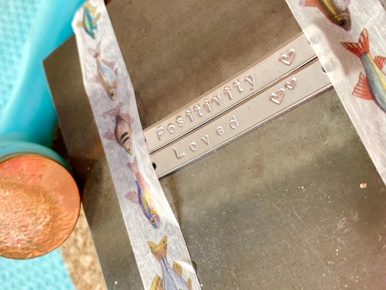K&ouml;nnte beinhalten: Zwei silberne Metallarmb&auml;nder mit den Worten "Positivity" und "Loved" sind abgebildet. Kleine Herzformen sind neben den Worten gestempelt. Dekoratives Klebeband mit bunten Fischmotiven ist auf dem Metall angebracht.