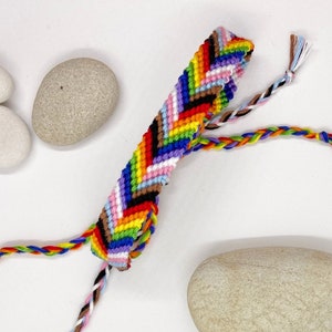 Puede incluir: Un brazalete de amistad multicolor con un patrón de chevron. El brazalete está hecho con hilos de diferentes colores, incluyendo rojo, naranja, amarillo, verde, azul, morado, rosa, marrón, negro y blanco.