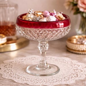 May include: A clear glass candy dish with a ruby red rim, filled with wrapped candies. The dish sits on a white lace doily. A decanter and flowers are in the background.
