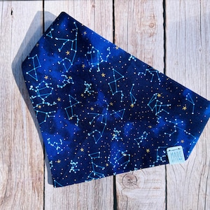May include: A blue fabric with a constellation pattern featuring white lines and gold stars. The fabric is folded and lies on a wooden surface. A small tag with the text "MADE WITH" is visible.