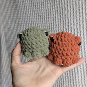 May include: Two small, round, crocheted plush figures made from thick, textured yarn. One figure is sage green and the other is burnt orange. Both have small, dark, circular eyes and rounded protrusions on the sides, resembling simple animal shapes.